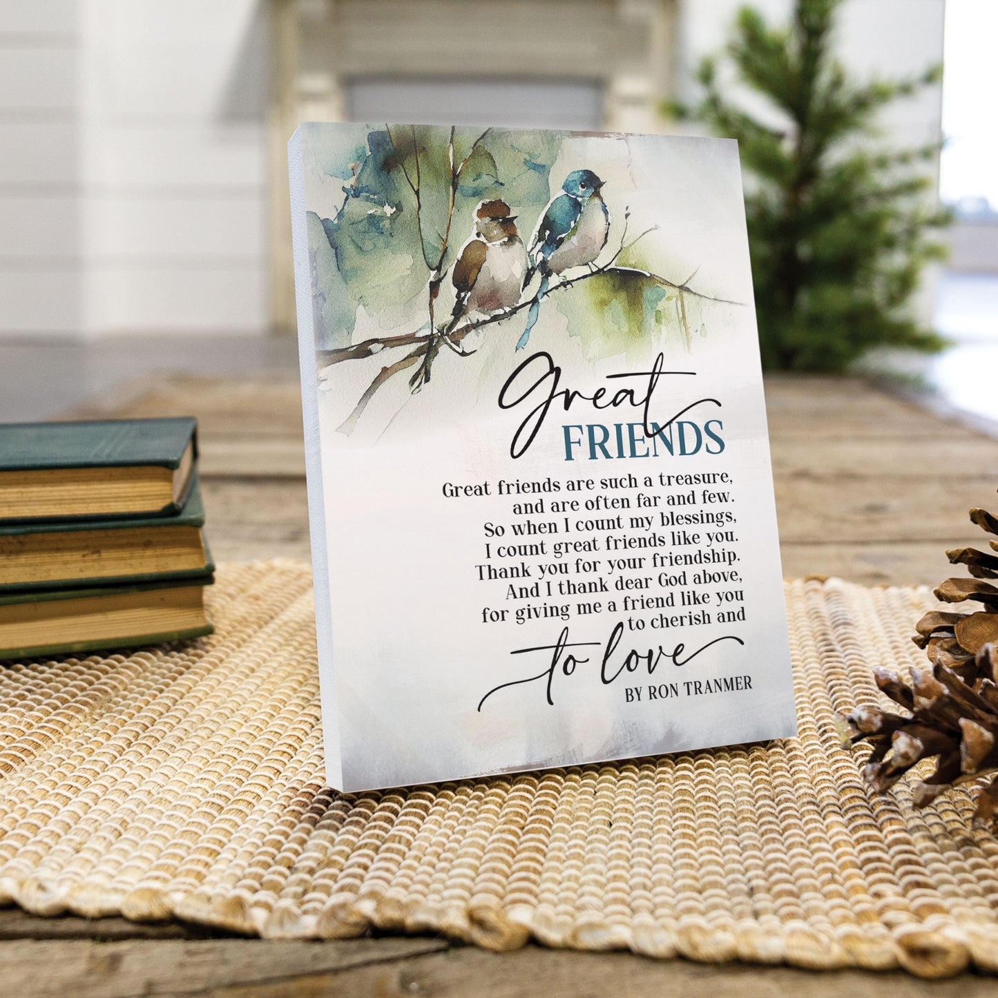 Decorative plaque with birds on a branch and text about great friends, placed on a woven mat with books and a pine cone. | oak7west.com