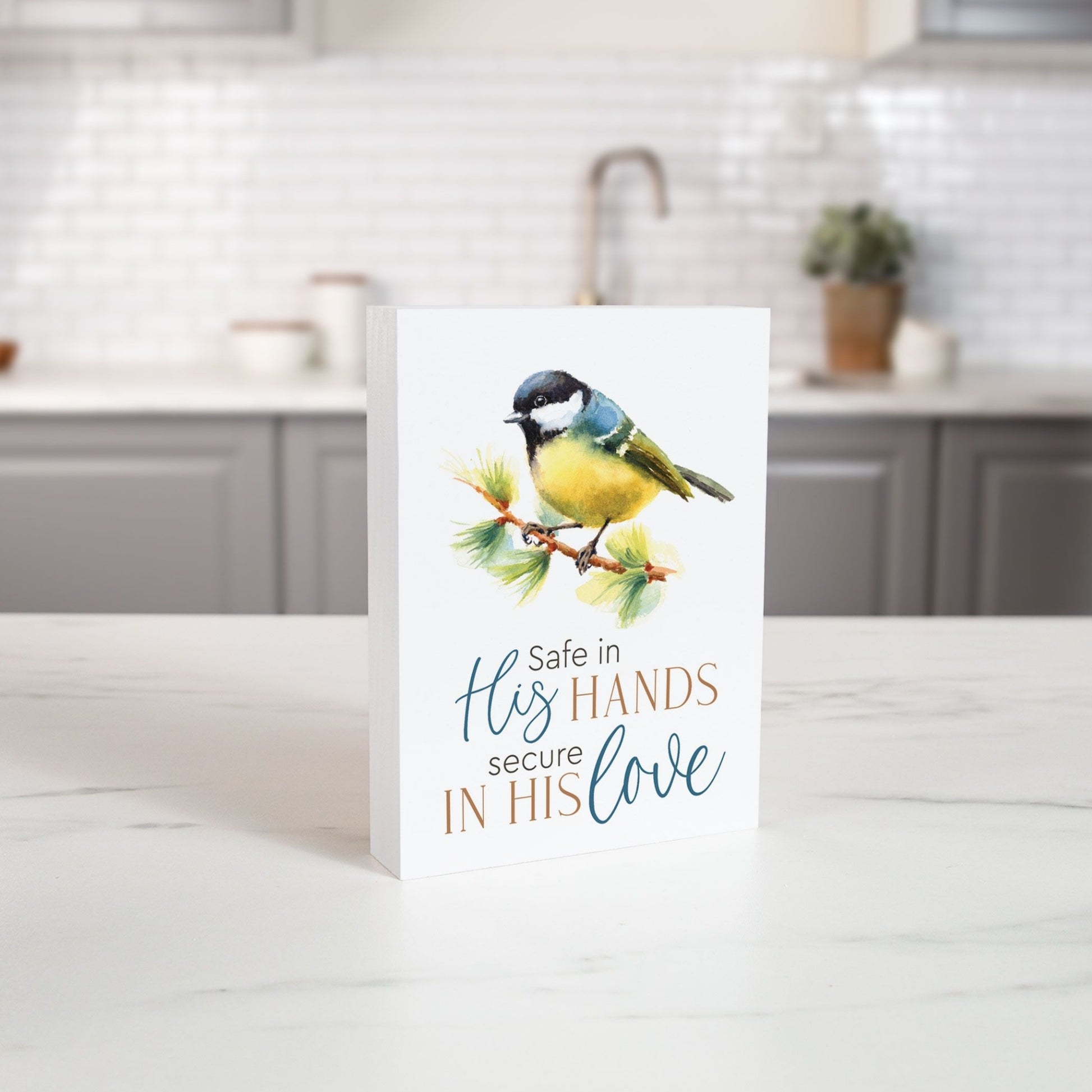Wood Block with a bird illustration and text 'Safe in His Hands, Secure in His Love' on a kitchen counter. | oak7west.com