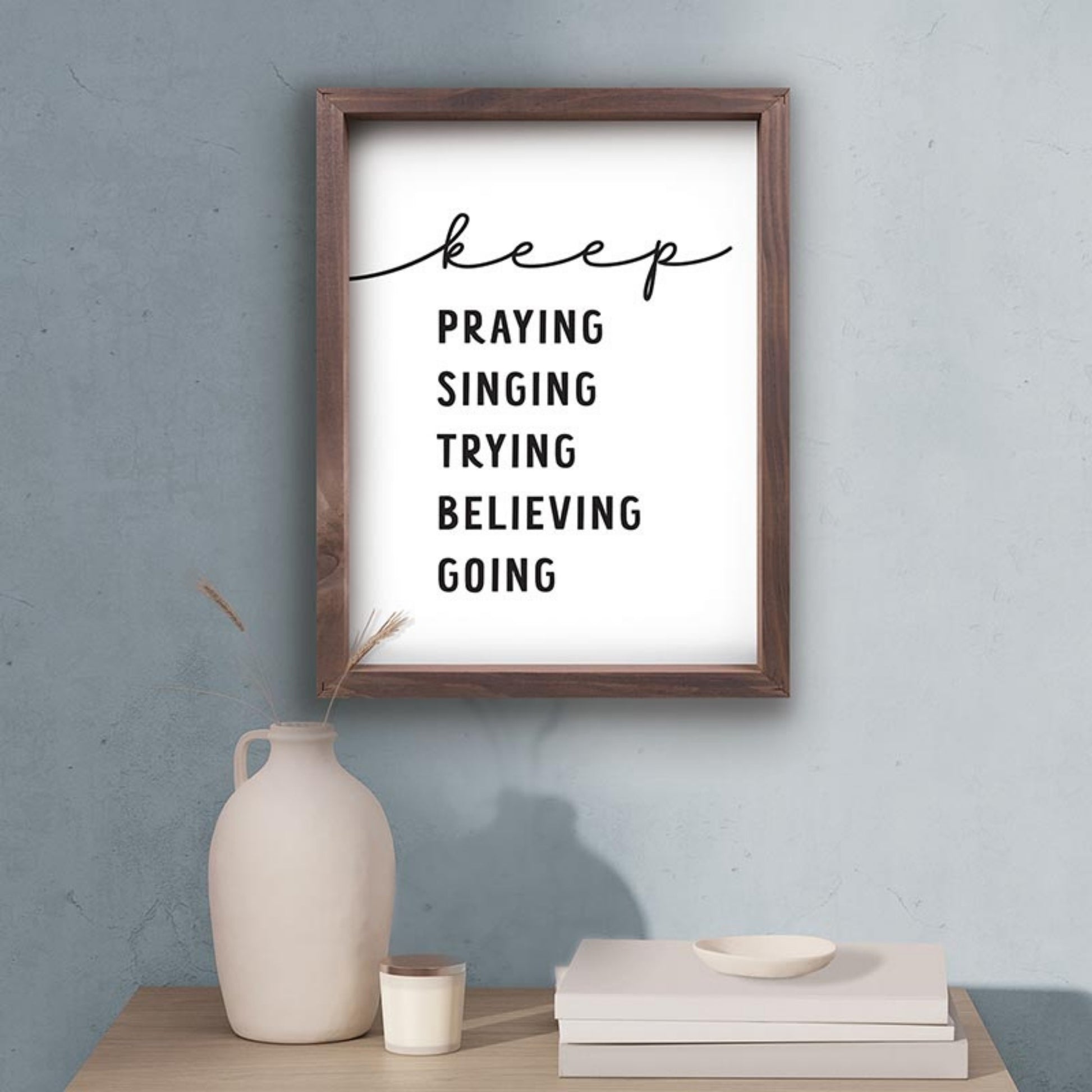 Framed motivational poster with inspirational quote on a wall above a table with decor items | keep PRAYING SINGING TRYING BELIEVING GOING - Inspirational Christian Wall Decor | oak7west.com